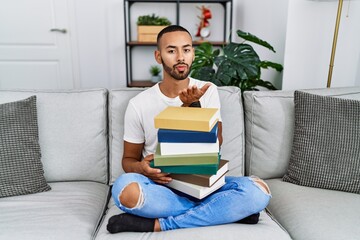 Sticker - African american young man holding a pile of books sitting on the sofa looking at the camera blowing a kiss with hand on air being lovely and sexy. love expression.