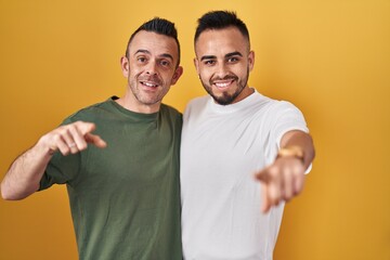 Sticker - Homosexual couple standing over yellow background pointing to you and the camera with fingers, smiling positive and cheerful