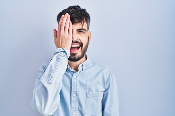 Wall Mural - Young hispanic man with beard standing over blue background covering one eye with hand, confident smile on face and surprise emotion.