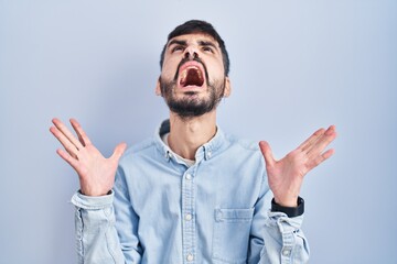 Wall Mural - Young hispanic man with beard standing over blue background crazy and mad shouting and yelling with aggressive expression and arms raised. frustration concept.