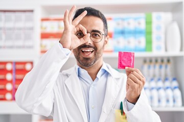 Sticker - Hispanic man with beard working at pharmacy drugstore holding condom smiling happy doing ok sign with hand on eye looking through fingers