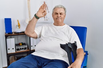 Canvas Print - Senior caucasian man at physiotherapy clinic holding crutches smiling positive doing ok sign with hand and fingers. successful expression.