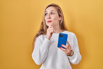 Canvas Print - Young blonde woman using smartphone typing message with hand on chin thinking about question, pensive expression. smiling and thoughtful face. doubt concept.