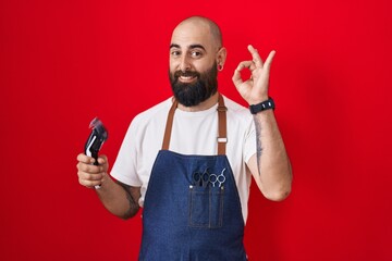 Canvas Print - Young hispanic man with beard and tattoos wearing barber apron holding razor smiling positive doing ok sign with hand and fingers. successful expression.