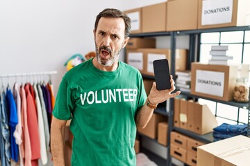 Canvas Print - Middle age man with beard wearing volunteer t shirt holding smartphone in shock face, looking skeptical and sarcastic, surprised with open mouth