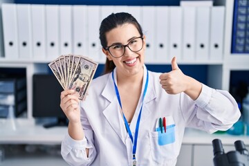 Canvas Print - Young brunette woman working at scientist laboratory holding money smiling happy and positive, thumb up doing excellent and approval sign