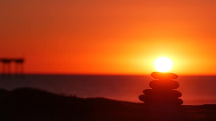 Wall Mural - Stack of pebble stones, sandy ocean beach, sunset sky. Rock balancing in sun light, sea water waves. Stones staking in pyramid pile. Zen meditation and harmony in balance. Seamless looped cinemagraph.