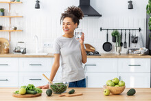 Woman Cooking Healthy Food With Glass Of Clean Water, Looking Away And Smiling. African American Young Female Preparing Fresh Salad For Dinner Or Breakfast In Home Kitchen. Healthy Diet Eating