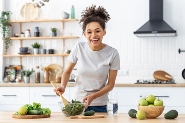 Canvas Print - Portrait of happy young african american woman cooking fresh salad for dinner or breakfast standing at home kitchen. Healthy lifestyle concept