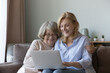 © fizkes - Mature daughter and old mother sit on sofa with laptop choose goods, buy services via internet, web surfing together at home. Younger relative teaching using computer, showing useful apps and websites