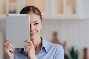 Adorable blonde young woman in blue shirt looks at camera covers her half face by tablet. Close up of confident businesswoman satisfied by technology and gadgets, against kitchen, mockup.