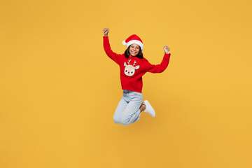 Merry fun little kid teen girl 13-14 years old wear red xmas sweater with deer Santa hat posing jump high doing winner gesture isolated on plain yellow background. Happy New Year 2023 holiday concept.