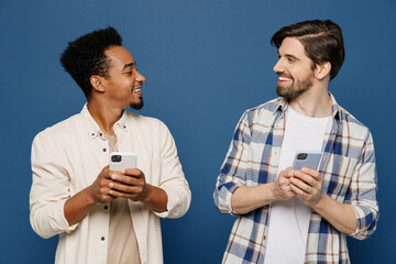 Wall Mural - Young two friends cheerful smiling happy cool men 20s wear white casual shirts together hold in hand use mobile cell phone chatting look to each other isolated plain dark royal navy blue background.
