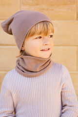 Wall Mural - a blond boy with dimples on his cheeks in a beige hat and a snood on the background of a stone wall