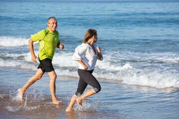 Mature couple running with joy along seashore. Happy Caucasian man and woman in sportswear training in summer and getting positive emotions of blue sea. Active life, health care of aged people concept