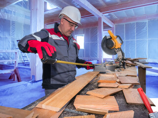 Poster - Man carpenter at work. Worker measures board with tape measure. Guy in gray work uniform. Man carpenter inside wooden house under construction. Carpenter career. Stationary circular saw on table