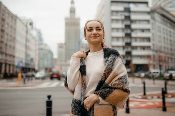 Happy young adult woman smiling with teeth smile outdoors and walking on city street weating winter clothes and knitted scarf. Female young traveler in Warsaw, Poland.