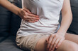 Close up of young female with fatty liver touches right side with hand, suffering from abdominal pain sit on grey couch. Pain in right side, appendix, gallstones and gynecological diseases concept