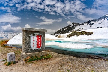 Sticker - Monument of Flag and coat of arms of the canton of Valais with snowy Col du Nufenen mountain