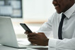 © Mediaphotos - Close-up of African American businessman using smartphone and smiling while sitting at table in front of laptop