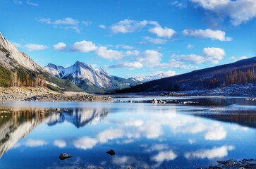 Canvas Print - Landscape with hills, snowy mountains and trees reflected in lake water under blue sky