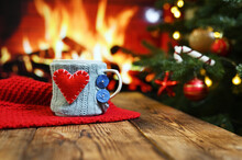 A Mug In A Knitted Case With A Red Heart On The Side On A Wooden Table Against The Background Of A Red Plaid, A Fireplace And A Christmas Tree