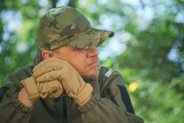 Poster - mature man dressed in camouflage uniform, green jacket, cap, tactical yellow leather-textile gloves sitting outdoor green forest and sky background. Tactical camouflage uniform clothes