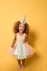 Funny child girl with party hat wearing a festive dress standing isolated over yellow background.