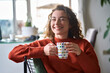 Young smiling pretty woman holding cup drinking warm tea or coffee relaxing dreaming at home. Happy positive lady enjoying hot drink daydreaming with mug in hands in cold cozy winter weekend morning.