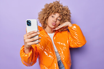 Wall Mural - Pretty curly haired stylish woman keeps lips folded poses at smartphone camera for making selfie has video conference photographs herself dressed in orange jacket isolated over purple background.