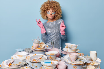 Household and home hygiene routine concept. Thoughtful woman wears festive silver dress rubber protective gloves surrounded by pile of dirty dishes with food leftovers poses indoor on blue background