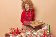 Happy woman glad to bake christmas cookies holds tray with pastries prepares for Xmas Eve going to decorate biscuits wears red jumper garland around neck looks positively aside beige background