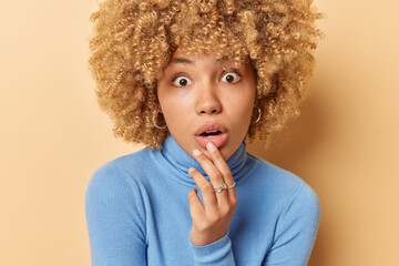 Poster - Headshot of amazed curly haired woman keeps mouth opened hand near lips stares with omg expression at camera wears blue turtleneck isolated over beige background cannot believe in shocking news