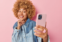 Fashionable Curly Haired Woman Dressed In Denim Jacket Clicks Selfie Via Smartphone Smiles Broadly Keeps Hand Under Chin Shows White Teeth Takes Photo Of Herself Isolated Over Pink Background.