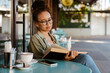 © Drobot Dean - White young woman drinking coffee while reading book in cafe outdoors