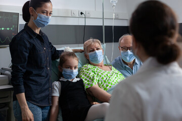 Wall Mural - Unrecognizable nurse explaining cares for elderly lady. Various ages people visiting old female patient hospital room. Family members, general practitioner wearing face masks at clinic rest room.