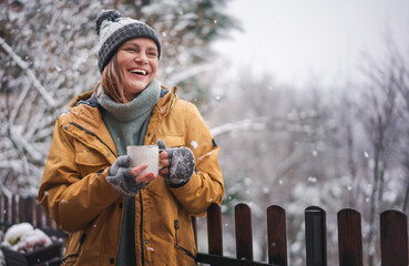 Young happy adult caucasian woman smiling in hat and yellow jacket enjoying tea in a mug with snowfall on the terrace