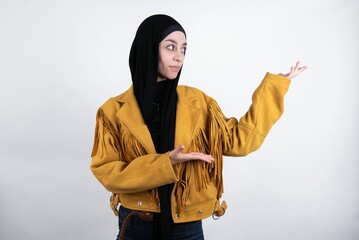 young beautiful muslim woman wearing hijab and yellow jacket over white wall pointing aside with both hands showing something strange and saying: I don't know what is this. Advertisement concept.