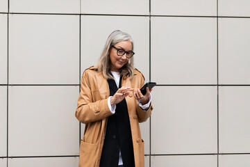 stylish businesswoman 50 years old with a mobile phone in her hands against the background of a beige wall outside