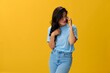 © SHOTPRIME STUDIO - Woman blogger with a phone in her hands in a blue t-shirt and jeans on a yellow background smile signs gestures symbols, online communication and video call, copy space, free background
