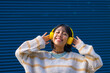 © unai - Asian girl listening to music in yellow headphones with her eyes closed on a blue college background