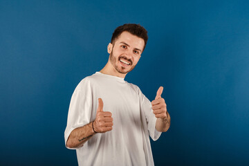 Wall Mural - Cheerful caucasian man wearing casual clothes posing isolated over blue background smiling and showing thumbs up at camera. Approving expression looking at the camera showing success.