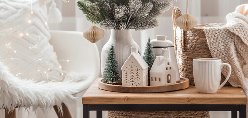 A bouquet of fir trees, a plaid in a wicker basket and Scandinavian white houses on a wooden table in the home interior of the living room. A cozy concept of festive home decoration.