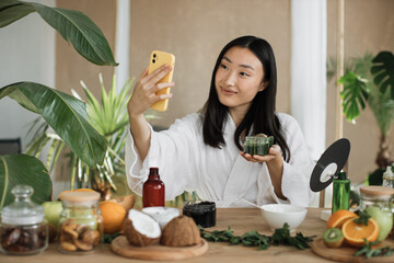 Asian brunette shoulder length hair female social media influencer in bathrobe streaming and introducing beauty products to audience with phone holding glass jar with cream, skin care concet.