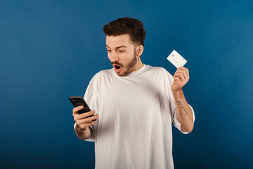 Wall Mural - Portrait of cheerful man wearing white t-shirt posing isolated over blue background holding smartphone and credit card shocked with surprise and amazed expression, fear and excited face.