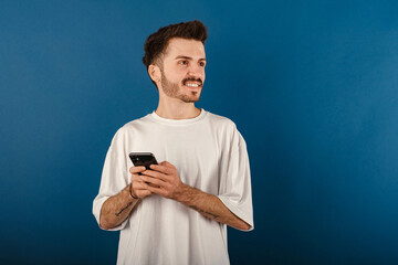 Wall Mural - Cheerful young man wearing t-shirt posing isolated over blue background dreamy look, thinking while holding smartphone and looking aside.