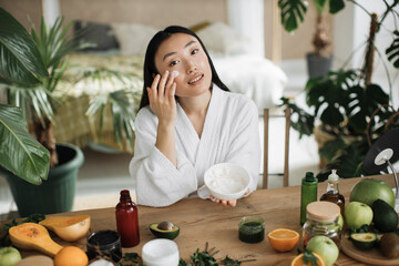 Beautiful asian woman preparing homemade cream with natural ingredients for skin care applying on face sitting at wooden table with various organic fresh vegetables and fruits on light exotic studio