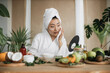 © sofiko14 - Healthy asian woman in white bathrobe and towel sitting at wooden table with various ingredients preparing natural cosmetics at home mixing mass in bowl while preparing cream and applying on face.