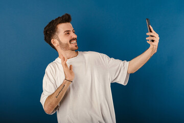 Wall Mural - Caucasian young man wearing t-shirt posing isolated over blue background taking selfie or having video call and waving hand. Recording vlog, social media influencer streaming, making video call.