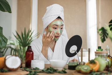 attractive asian woman looking at mirror sitting at table with ingredients for homemade cosmetics ap